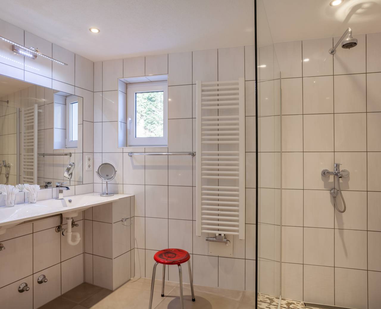 Weiß gefliestes Badezimmer mit Dusche, Waschbecken, Wandspiegel und Handtuchtrockner