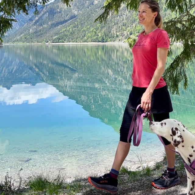 Eine Frau wandert mit ihrem Hund am türkisblauen Plansee entlang