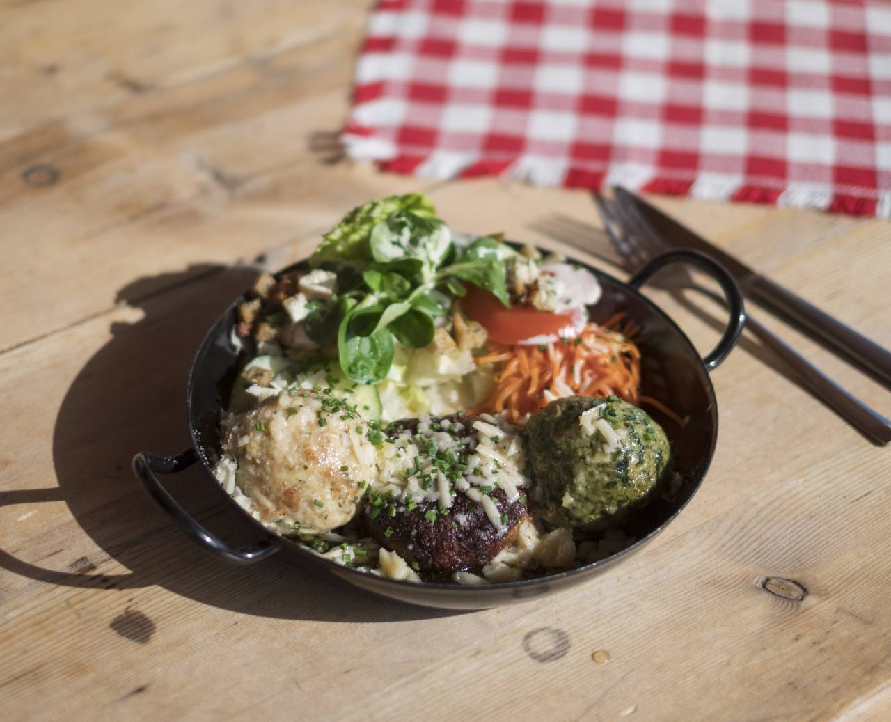Pfanne mit Knödel und Beilagensalat auf einem Holztisch mit Besteck und rotkarierter Tischdecke