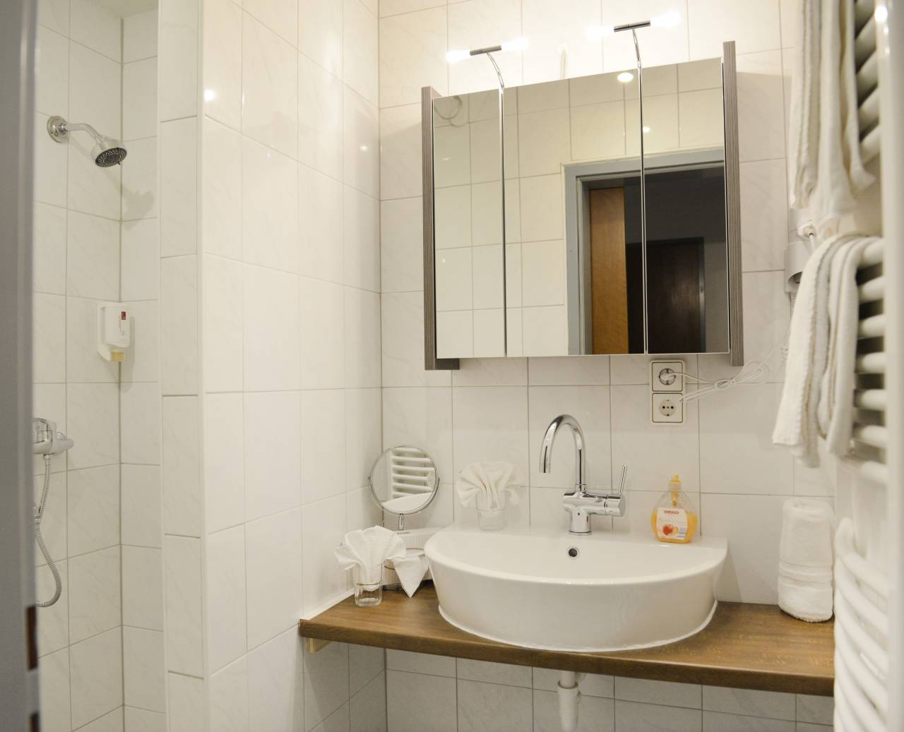 Weiß gefliestes Badezimmer mit Dusche, Waschbecken, Spiegelkasten und Handtuchtrockner